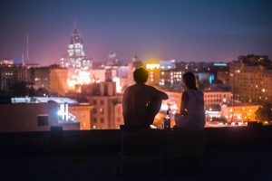 Two people sit on rooftop, looking out at city