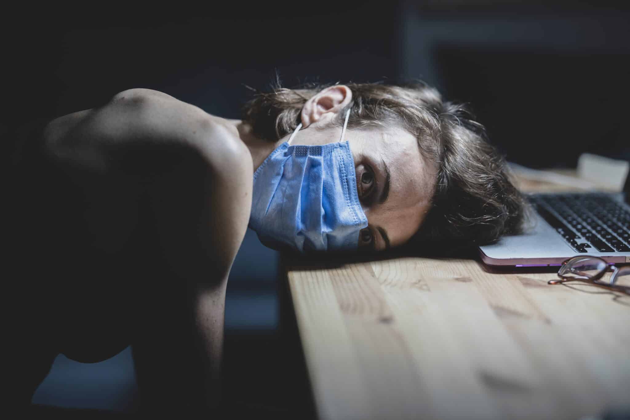 Woman in medical mask resting head on table near laptop, looking depressed.