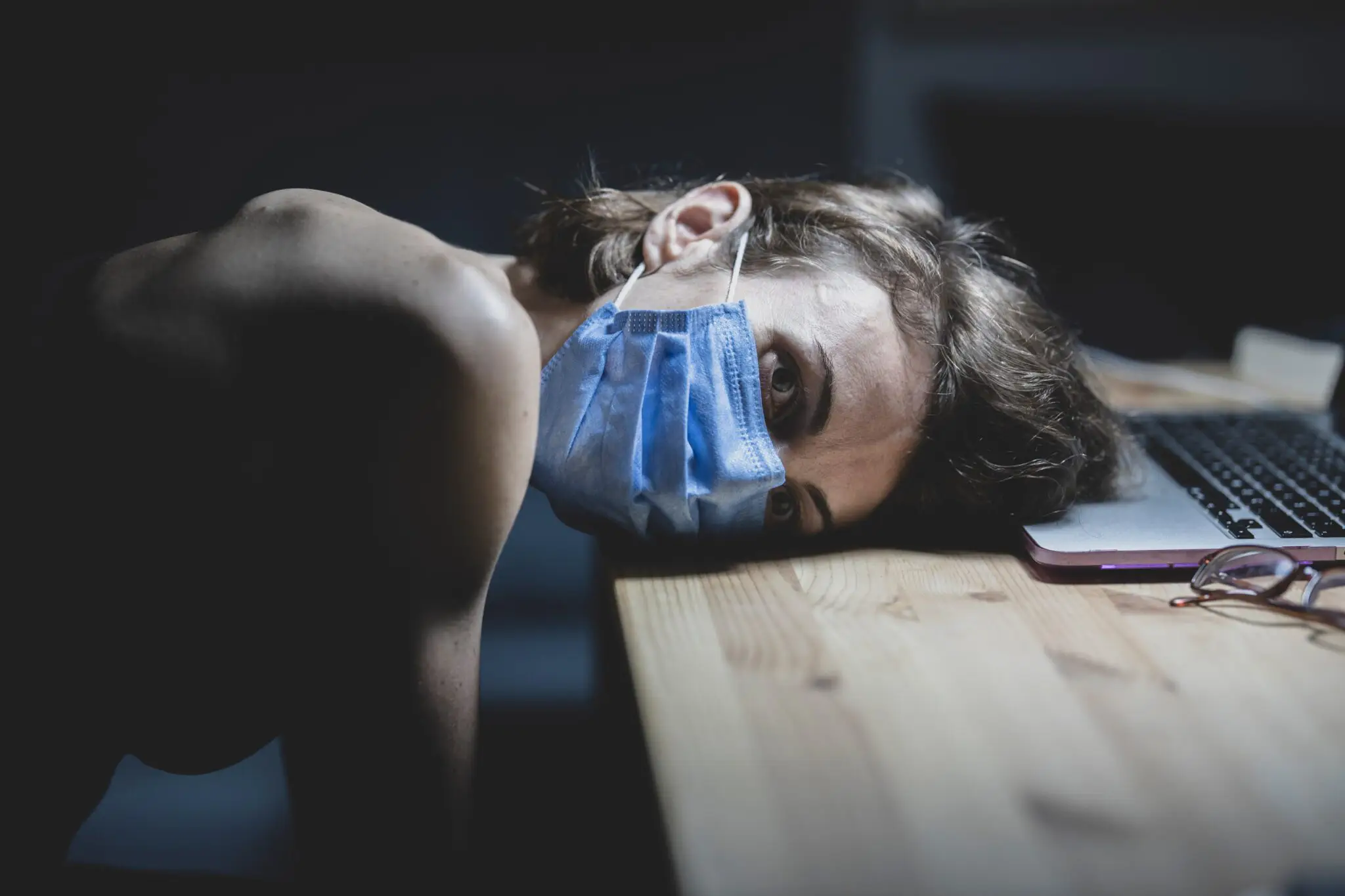 Woman in medical mask resting head on table near laptop, looking depressed.