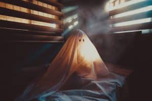 A ghost in a sheet sitting at the edge of a bed in the dark