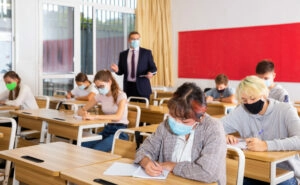 Focused teenage students in protective face masks studying in classroom with teacher, writing lectures in workbooks. Necessary precautions in coronavirus pandemic