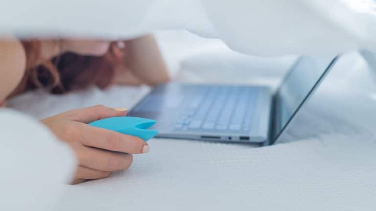 A woman lies in bed holding a clitoral vibrator and watching porn on a laptop