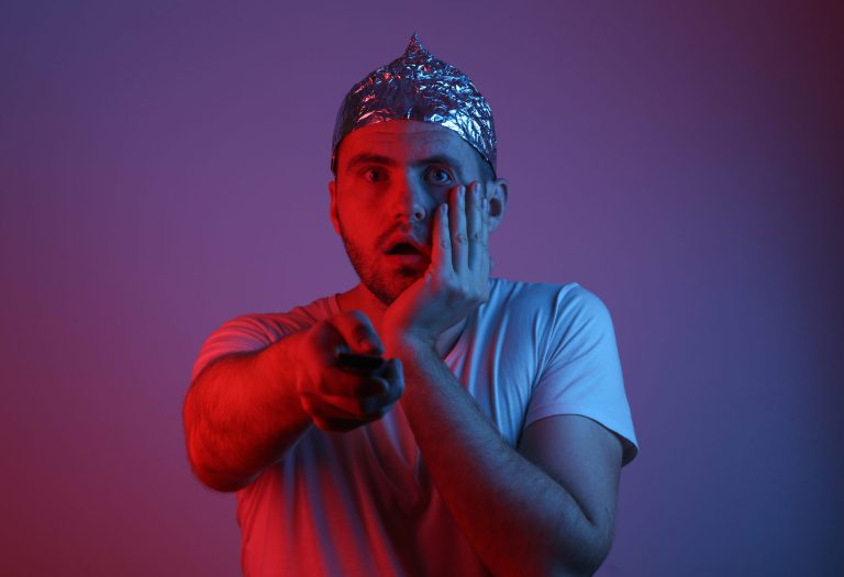 Surprised Bearded Man in a foil hat holds tv remote in red-blue neon light. Conspiracy