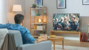 In the Living Room: Guy Relaxing on a Couch Watching War Movie on a TV. Modern Military Warfare Action with War Soldiers Shown on a Television.